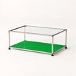 A modern modular coffee table with a clear glass top, a bright green lower shelf, and a stainless metal frame with black feet, shown on a clean white background.
