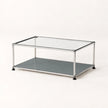 A modern two-tier coffee table with a clear glass top, a dark gray lower shelf, and a stainless chrome metal frame with black feet, shown on a clean white background.
