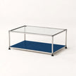 A modern modular coffee table with a clear glass top, a blue lower shelf, and a stainless metal frame with black feet, shown on a clean white background.