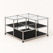 A modular two-tier storage organizer made of a chrome metal frame with black metal shelves and clear glass top panels, shown on a light cream background.