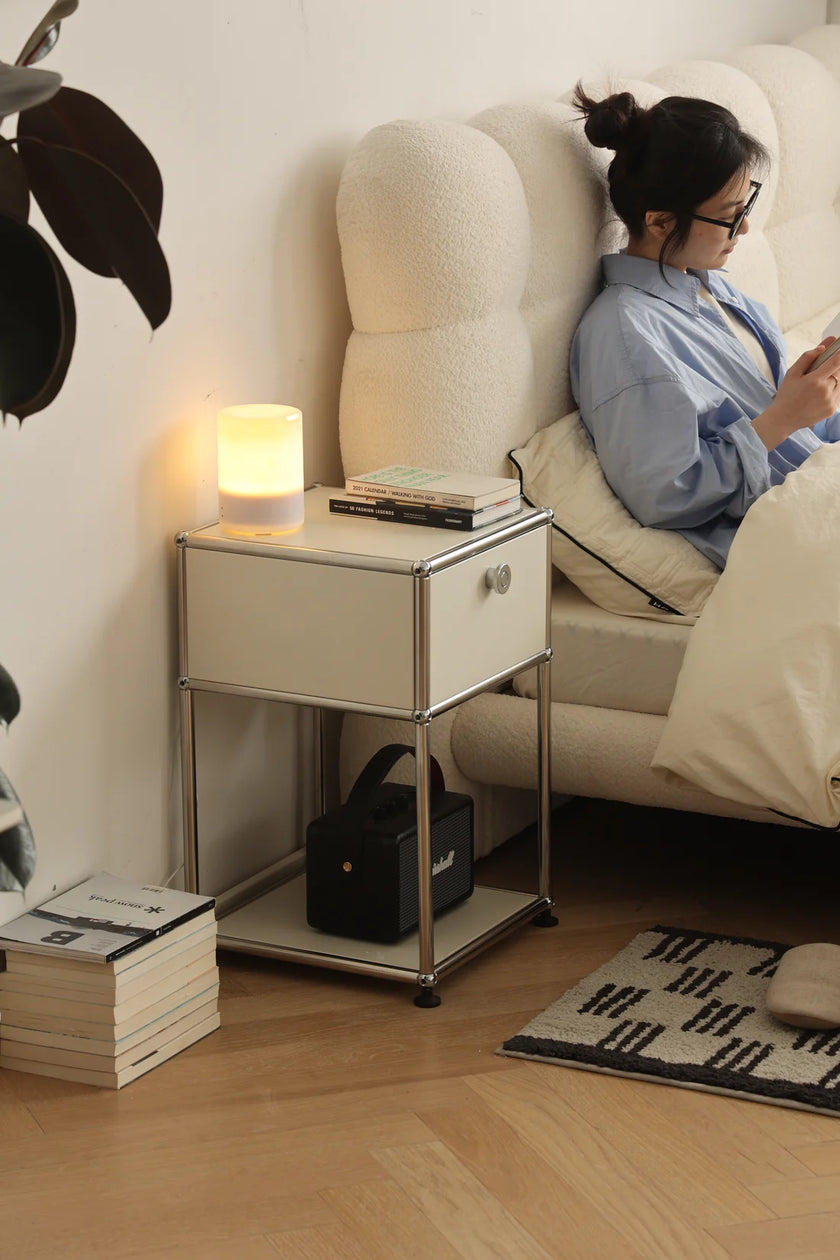 Lady lying in bed using a smartphone next to a inspired recreation USM modular nightstand with a lamp and books.