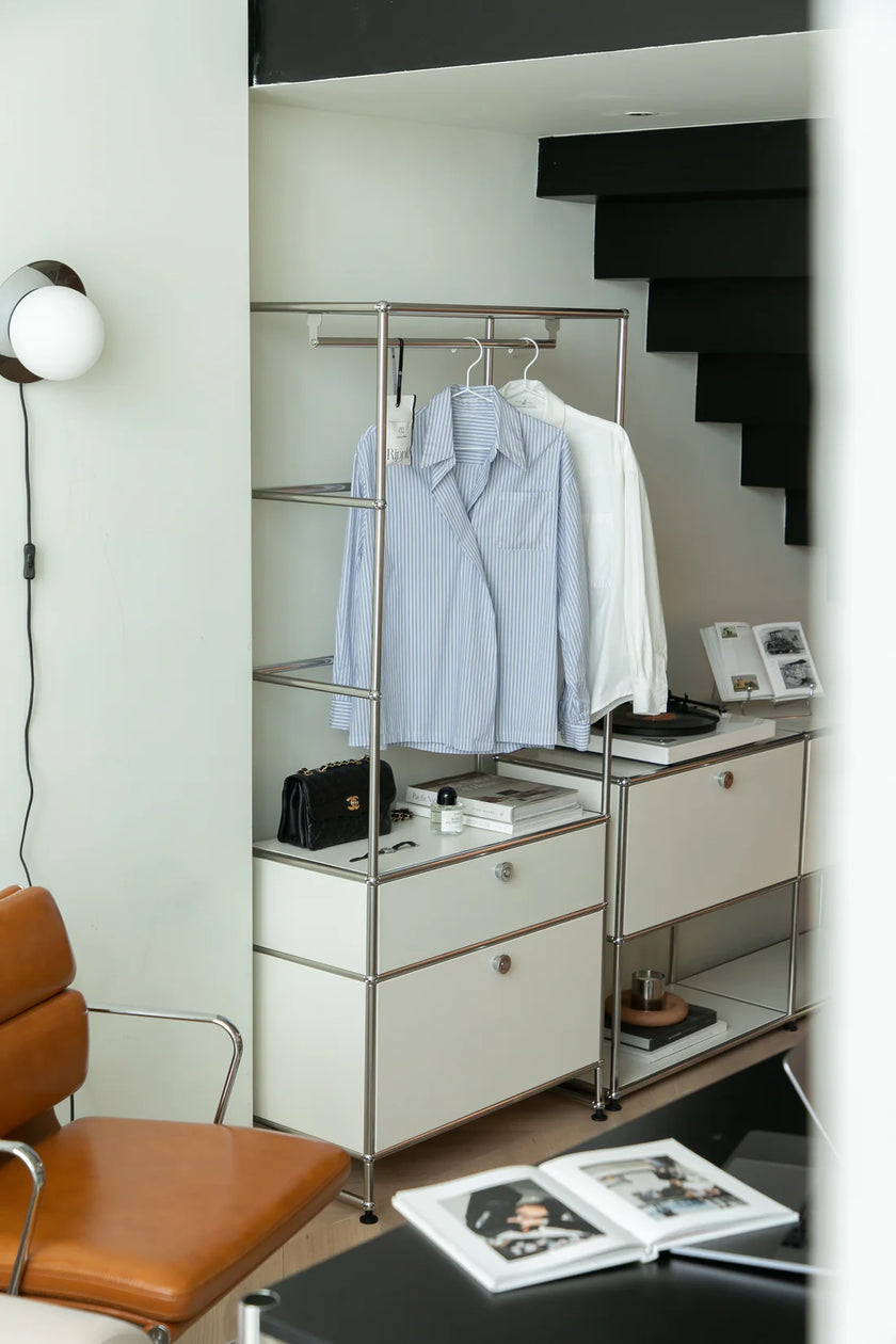 Closet with clothes on a inspired recreation USM modular rack, shelves, and a chair in a room.