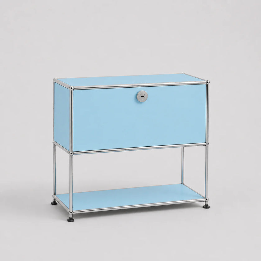 A light blue modular storage cabinet with a chrome metal frame, featuring a single drawer with a round silver knob and an open lower shelf, set against a neutral light gray background.