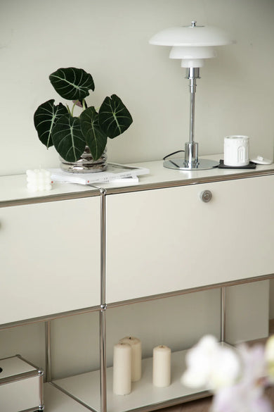 White inspired recreation USM modular cabinet with a plant, lamp, and candles on top against a light wall.