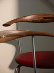 Close-up of a modern wooden chair with curved dark wood backrests, a red upholstered seat, and thin metallic legs against a neutral gray wall.