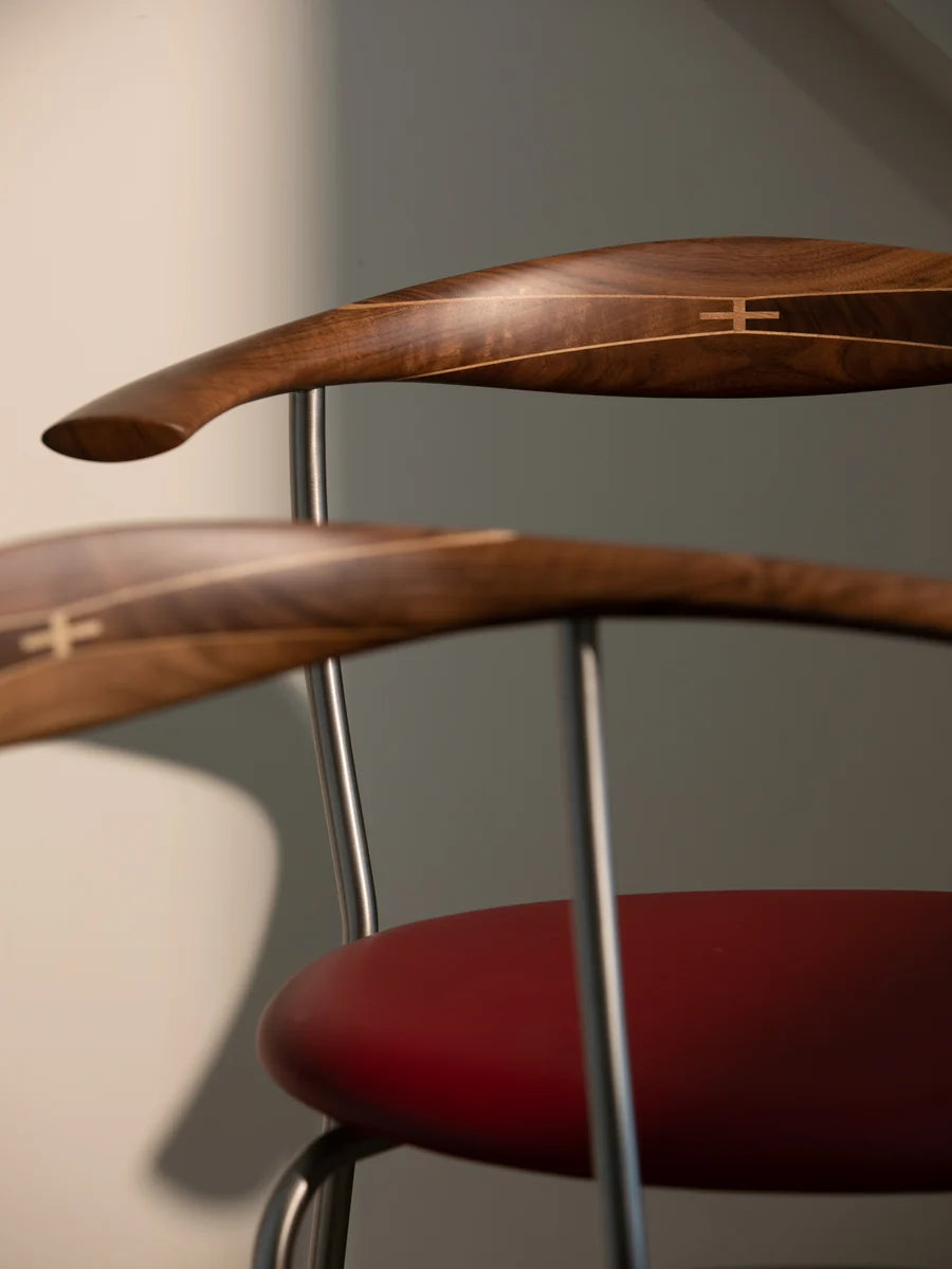 Close-up of a modern wooden chair with curved dark wood backrests, a red upholstered seat, and thin metallic legs against a neutral gray wall.