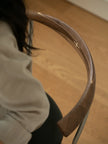 Close-up of a curved wooden chair back with a light wood inlay, shown in a real indoor setting with a wooden floor and part of a seated person visible on the left.