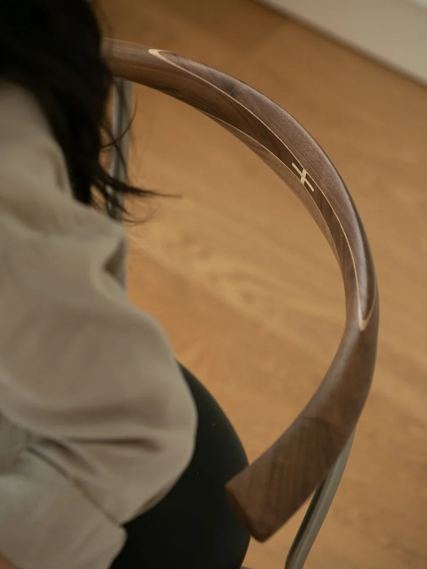 Close-up of a curved wooden chair back with a light wood inlay, shown in a real indoor setting with a wooden floor and part of a seated person visible on the left.
