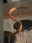A close-up of a modern wooden chair with a curved, spiral-shaped brown wood backrest and black seat, set in an indoor environment with a patterned rug and neutral background.