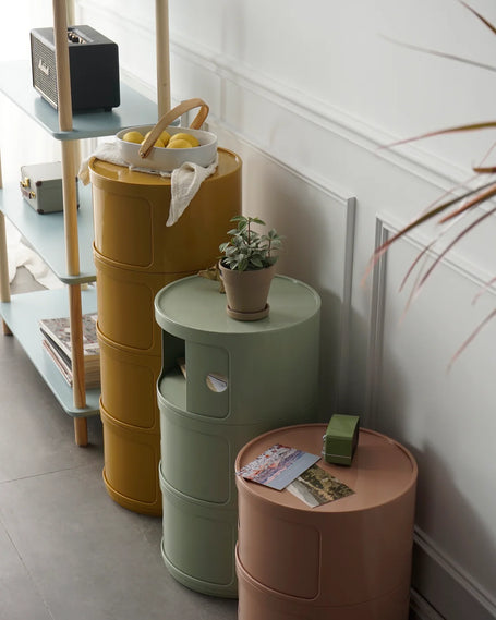 Three cylindrical plastic storage units in yellow, green, and pink against a white wall.