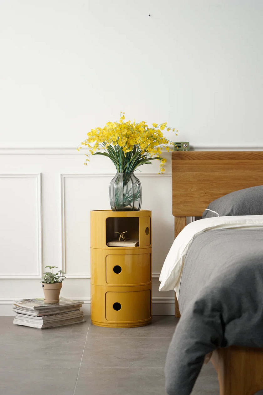 Yellow plastic storage containers nightstand with a vase of yellow flowers next to a bed in a bedroom.