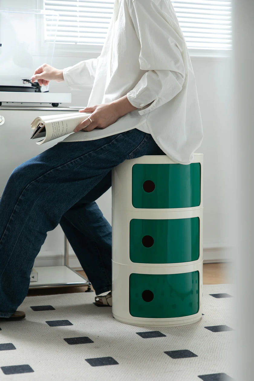 Lady sitting on a green and white storage units​ in a room with a desk and window blinds.