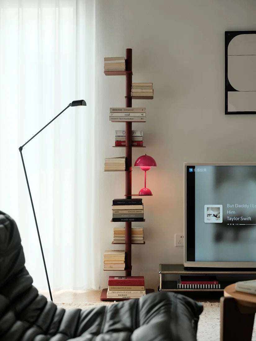 A tall, narrow bookshelf styled like a tree trunk with staggered horizontal book shelves, holding numerous books and a small pink lamp. It is set in a modern living room next to a black floor lamp, a black leather chair, and a television.