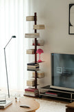 A tall, narrow dark wood bookshelf with floating shelves styled with books, a pink lamp, and decorative objects, set in a modern living room next to a TV stand and television.