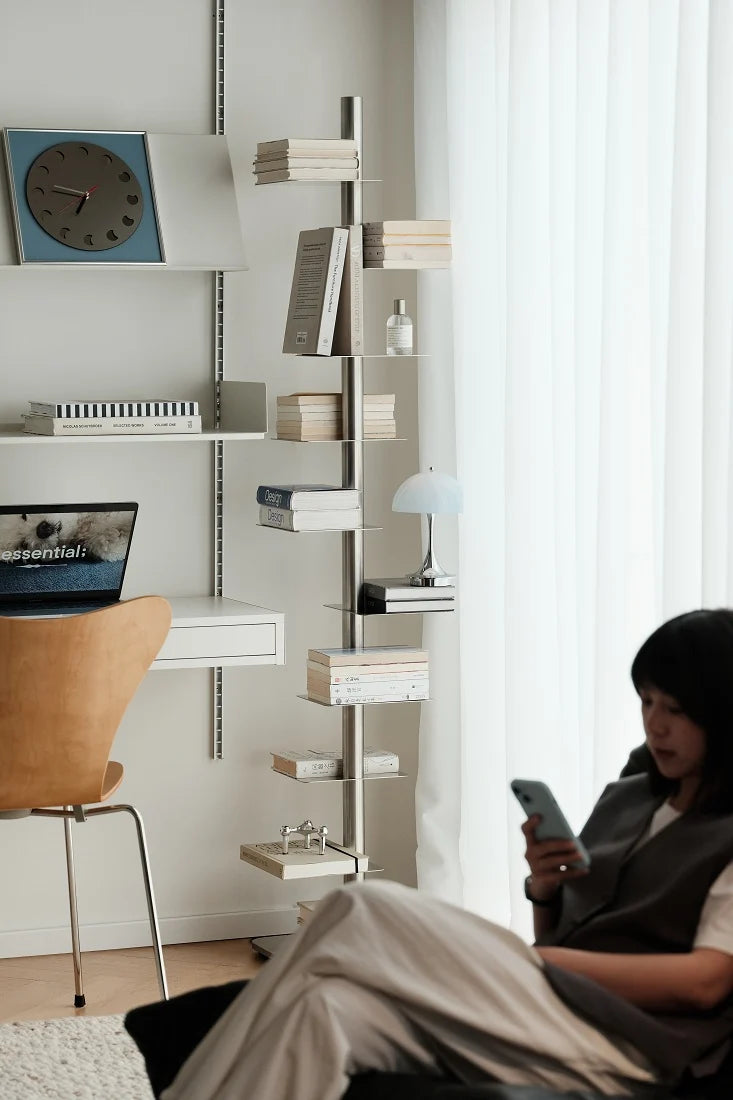A modern interior scene with a tall slim shelving unit holding books and small decor items, next to a desk with a chair and a laptop. A person reclines in the foreground near a window with sheer curtains, looking at a phone.