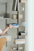 A modern wall-mounted shelving unit with a vertical metal frame and floating glass shelves, styled with books, a blue dome lamp, and accessories. A person's arm is visible reaching for a book on one of the shelves.