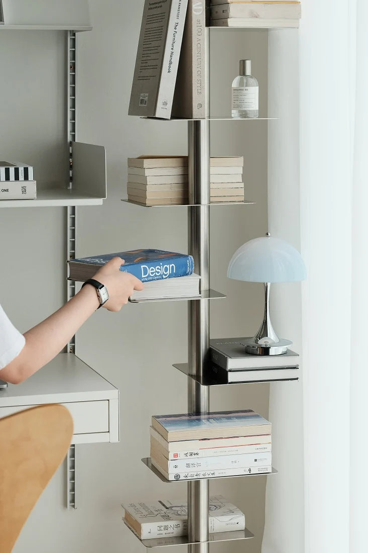 A modern wall-mounted shelving unit with a vertical metal frame and floating glass shelves, styled with books, a blue dome lamp, and accessories. A person's arm is visible reaching for a book on one of the shelves.