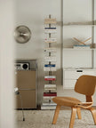 A tall, narrow metal bookshelf filled with books stands in a minimalist room next to a wooden chair, white shelving, and a small cabinet.