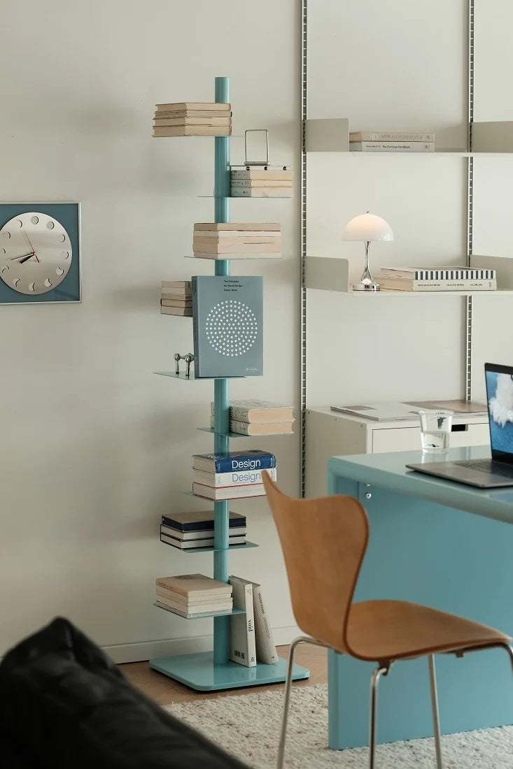 A tall, slender turquoise bookshelf with multiple tiers of open shelves and stacked books, styled in a modern home office next to a light blue desk and a brown leather chair.