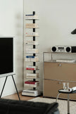 A tall, narrow black shelving unit with multiple horizontal glass shelves, styled with books in a modern living room setting next to a beige cabinet and a television.