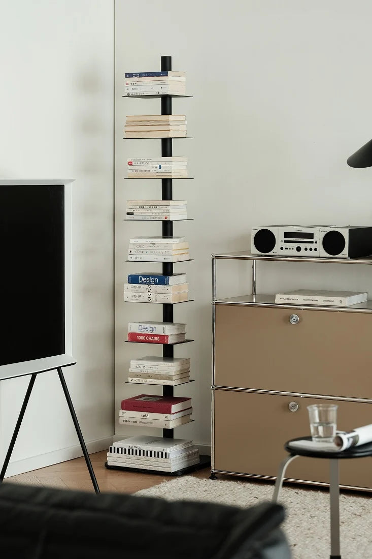 A tall, narrow black shelving unit with multiple horizontal glass shelves, styled with books in a modern living room setting next to a beige cabinet and a television.
