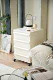 White nightstand with a lamp and books next to a bed in a bedroom setting.