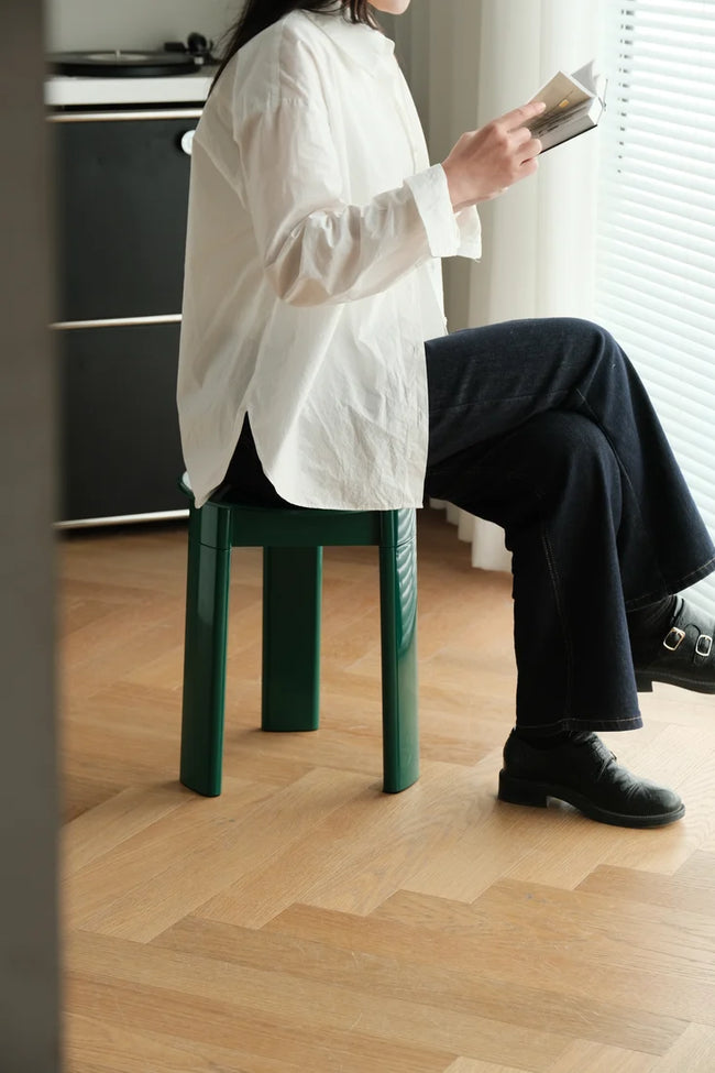 A person seated on a dark green stool in a bright room, wearing an oversized white shirt, dark wide-leg pants, and black shoes while holding a book.