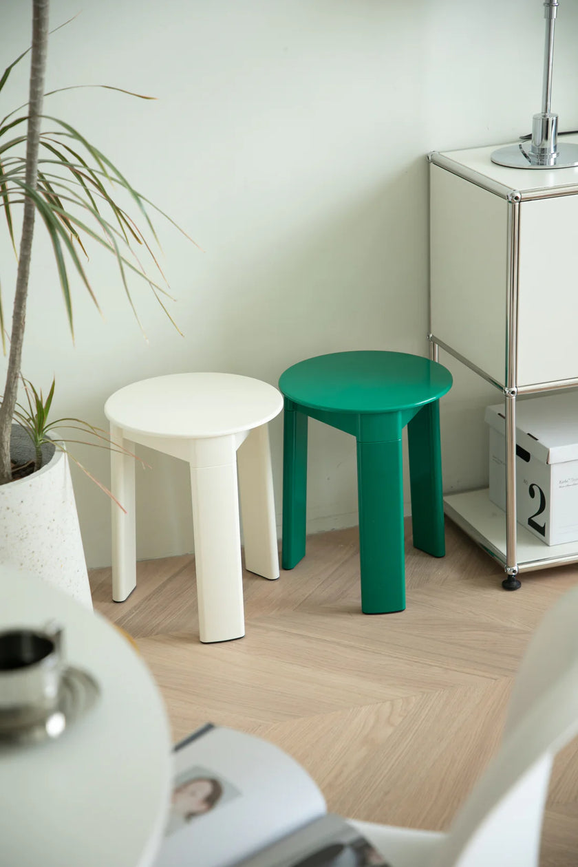 Two bathroom stools, one white and one green, on a wooden floor with a plant and cabinet in the background.