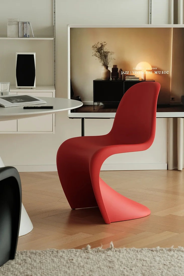A modern red molded plastic chair with a sculptural curved back and base, positioned in a minimalist, contemporary living room setting with light wood flooring and a white wall.