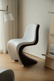 A modern sculptural chair with a white shell seat and back layered over a dark brown curved base, placed in a minimalist room with light wood flooring, a wall lamp, and shelving.