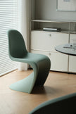 A modern sculptural chair in a muted sage green color, positioned in a minimalist room with light wood floors, a white cabinet, and a round table.