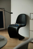 A modern sculptural black chair in a minimalist room with light wood flooring, a white wall, and a black floor lamp.