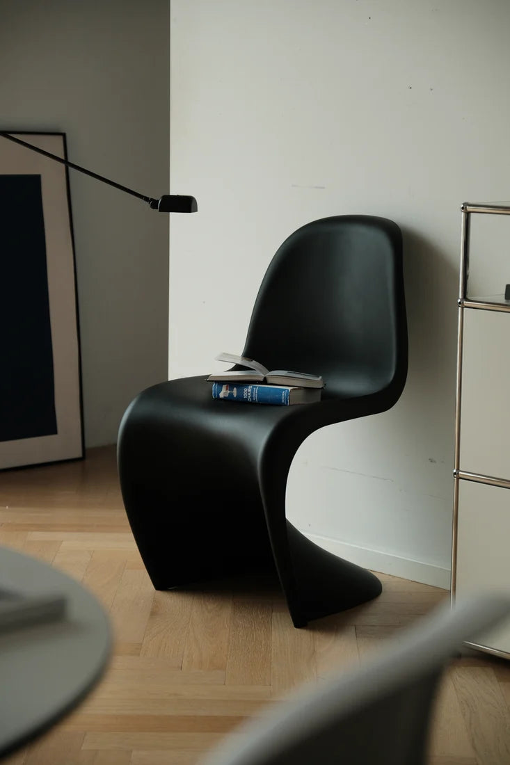 A modern sculptural black chair in a minimalist room with light wood flooring, a white wall, and a black floor lamp.