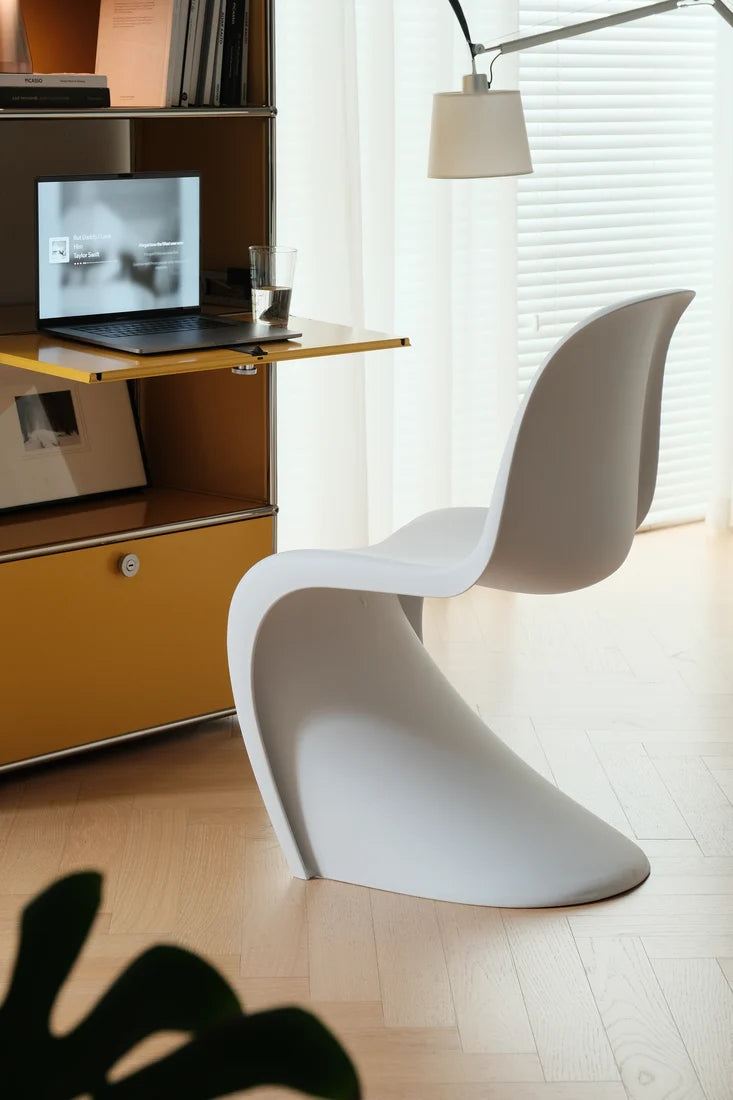 A modern white molded plastic chair in a bright, minimalist home office setting next to a wooden desk with a laptop and a floor lamp.