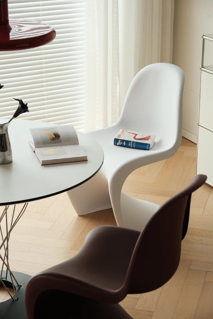 A modern white molded chair in a bright room next to a round white table, with a dark brown chair in the foreground and a window with blinds in the background.