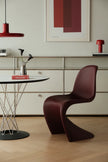 A modern sculptural chair in deep burgundy leather, positioned in a minimalist dining room setting with a round table, a red pendant light, and a cream-colored cabinet in the background.