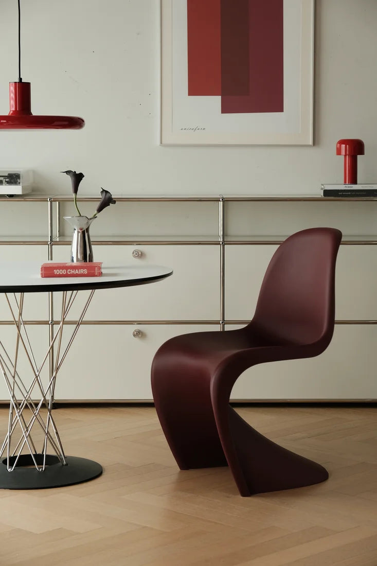 A modern sculptural chair in deep burgundy leather, positioned in a minimalist dining room setting with a round table, a red pendant light, and a cream-colored cabinet in the background.