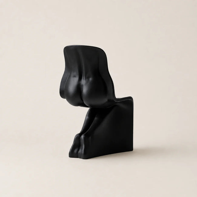 A minimalist black sculptural chair with a curved, abstract seat and back, set against a solid, warm off-white background.