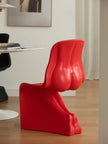 A bold, sculptural red chair with a highly curved, organic silhouette, shown in a modern room setting with light wood floors and a white wall.