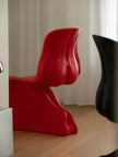 A bold, sculptural red chair with a glossy finish, positioned on a light wood floor in front of white curtains. A blurred black chair is visible in the foreground on the right.