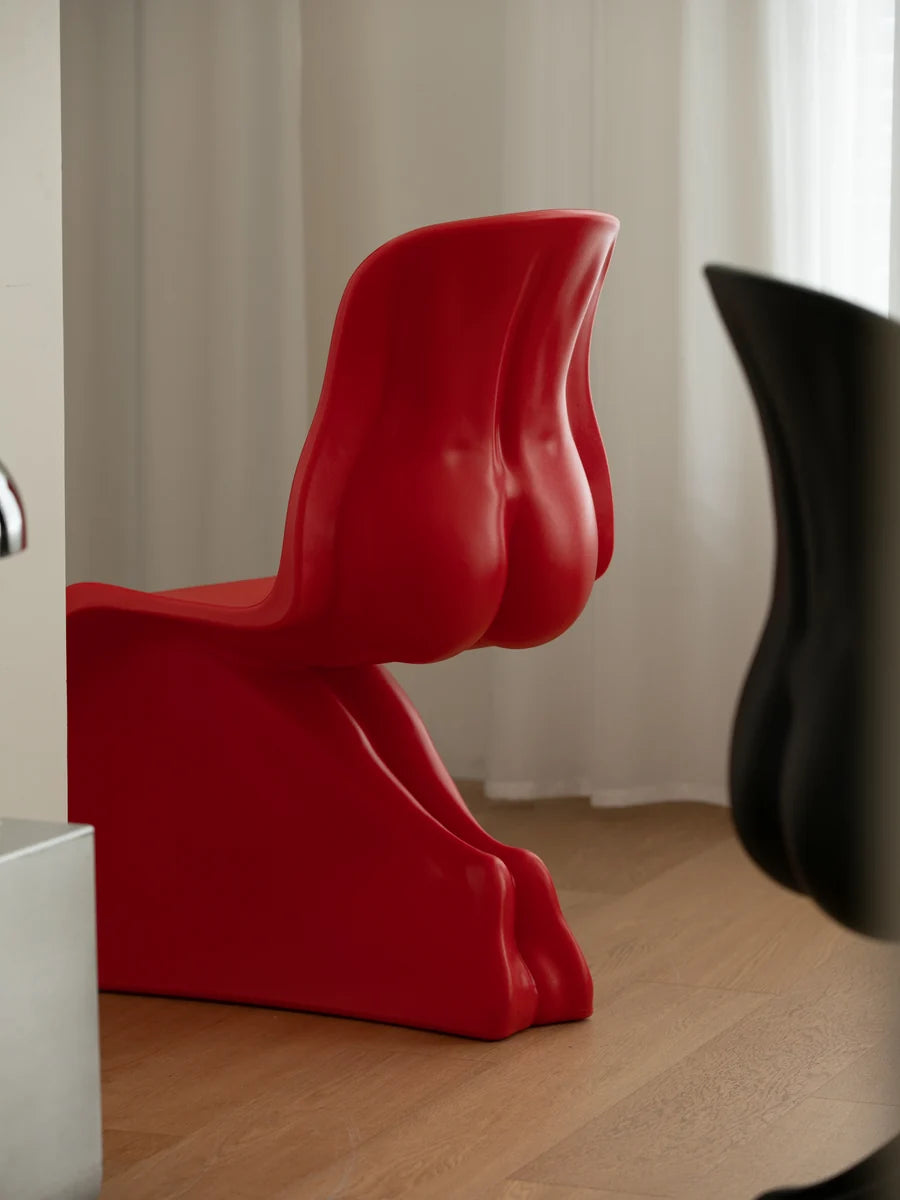 A bold, sculptural red chair with a glossy finish, positioned on a light wood floor in front of white curtains. A blurred black chair is visible in the foreground on the right.