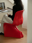 A woman sits on a bold, sculptural red molded chair in a modern indoor setting with light wood floors, a white table, and a laptop in the background.
