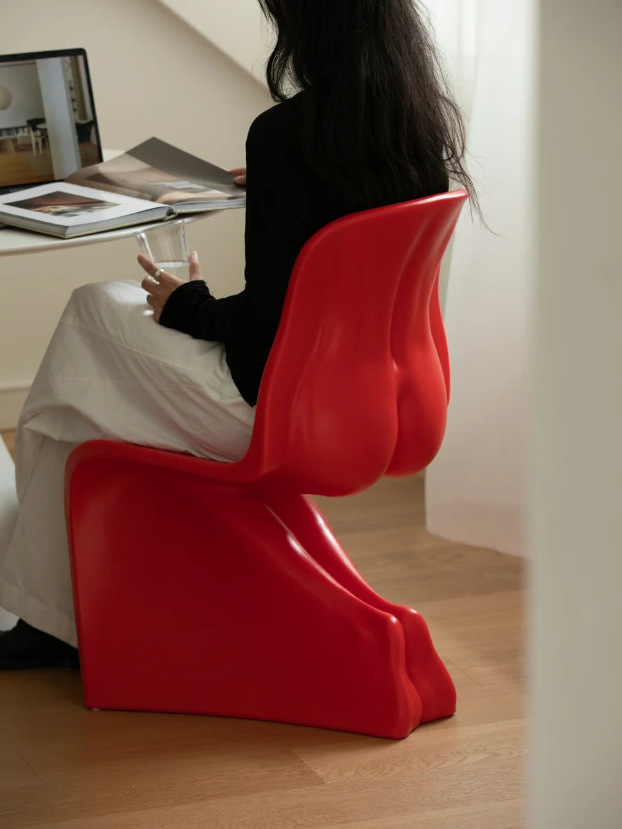 A woman sits on a bold, sculptural red molded chair in a modern indoor setting with light wood floors, a white table, and a laptop in the background.
