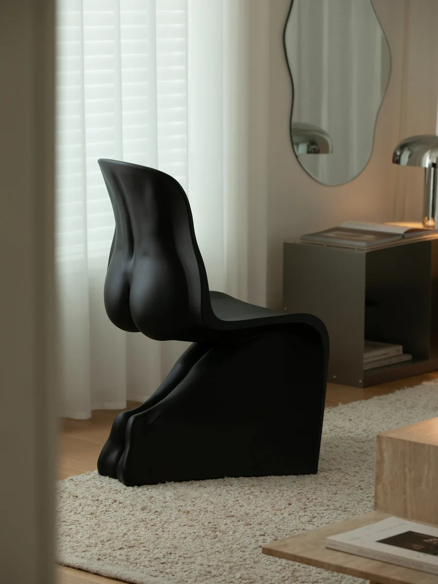 A sculptural black molded chair placed on a light beige rug in a modern interior room with curtains, a side table, and a mirror visible in the background.
