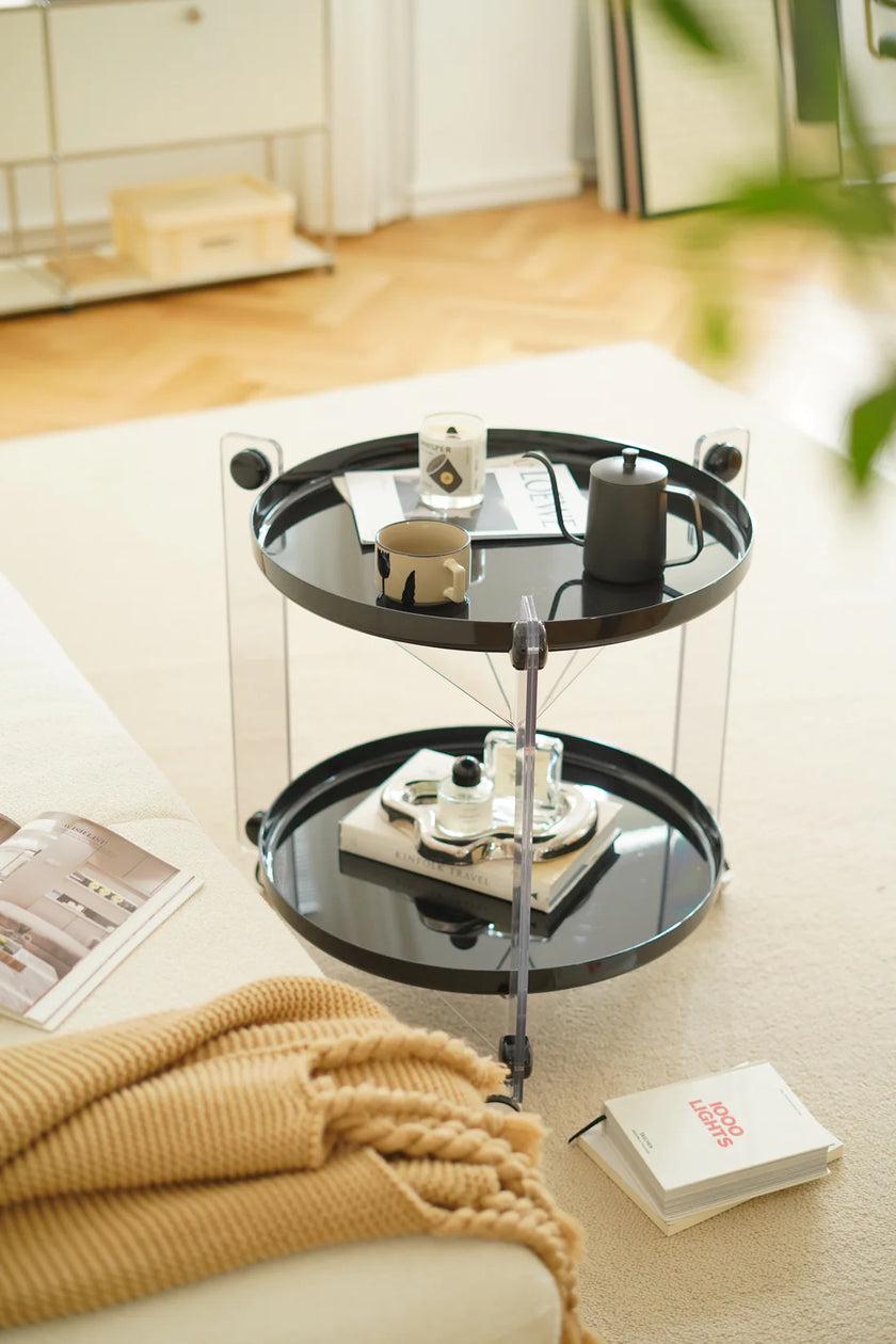 Round black side table with two tiers on a light-colored floor, surrounded by books and a blanket.