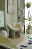Modern interior with a white fur chair cushions, plant, and abstract art on a light-colored floor.