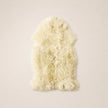A fluffy sheepskin rug in a warm cream or ivory color, laid flat against a solid, light beige background.