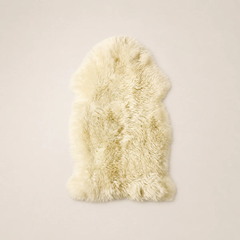 A fluffy sheepskin rug in a warm cream or ivory color, laid flat against a solid, light beige background.