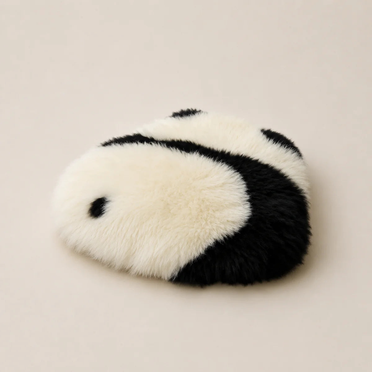 A plush faux-fur accessory with a black and white panda-inspired pattern, photographed on a plain warm beige background.