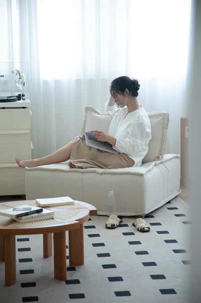 Woman sitting on a beige small sectional sofa using a laptop in a bright room with white curtains.